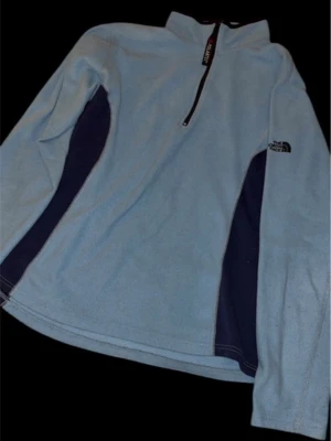Ljusblå fleece-tröja från The North Face - Säljer en ljusblå fleece-tröja från The North Face med mörkblå paneler på sidorna. Tröjan har lång ärm, half zip och hög krage. Perfekt för kyliga dagar och outdoor-aktiviteter. Polartec-material för extra värme och komfort.