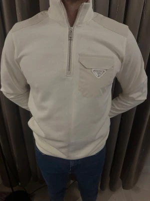 Vit half zip tröja från Prada - Säljer en stilren vit tröja från Prada med half zip och hög krage. Tröjan har en ficka med lock och metallmärke på bröstet. Materialet är mjukt och känns lyxigt, perfekt för dig som gillar exklusiva märken och clean look.
