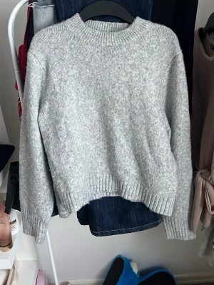 Grå stickad tröja med rund hals - Mysig grå stickad tröja med rund hals och ribbade muddar vid ärmslut och nederkant. Perfekt för kyliga dagar och enkel att matcha med jeans eller kjol. Tröjan har en normal passform och långa ärmar, vilket gör den både bekväm och stilren.