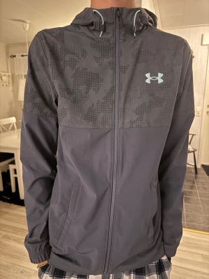 Under Armour grå vindjacka med camomönster - Snygg grå vindjacka från Under Armour med camomönstrad överdel och huva. Jackan har dragkedja framtill, två sidofickor och elastiska muddar. Materialet är lätt och syntetiskt, 