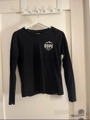 Svart långärmad t-shirt från Dope - Svart långärmad t shirt från Dope med vit logga och texten 'Paradise isn't tropical' på bröstet. Klassisk rund halsringning och liten tygpatch med bergmotiv vid nederkanten. Skön och enkel stil som funkar till det mesta. Lite slitage i trycket på ryggen
