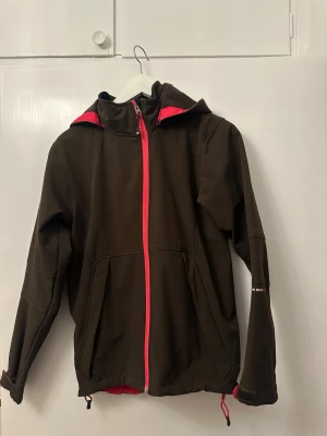 Brun vindjacka från North Bend - Säljer en brun vindjacka från North Bend med huva och snygga röda detaljer. Jackan har dragkedja framtill, två fickor och justerbar nederkant. Insidan är fodrad med mjuk fleece för extra komfort. Perfekt för blåsiga dagar och outdoor-häng.