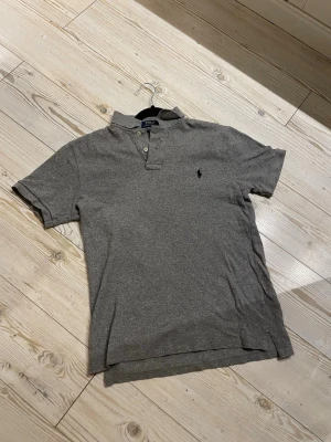 Grå pikétröja Polo Ralph Lauren M - Klassisk grå pikétröja från Polo Ralph Lauren i slim fit-modell. Tröjan har korta ärmar, krage med två knappar och den ikoniska broderade loggan på bröstet. Tillverkad i mjuk bomull som är skön mot huden.