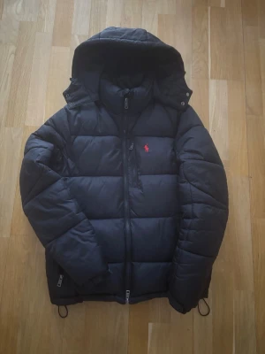 Svart pufferjacka från Polo Ralph Lauren - Svart pufferjacka från Polo Ralph Lauren med huva och dragkedja framtill. Jackan har ett rött broderat Polo-logo på bröstet och praktiska fickor med dragkedja. Perfekt för kalla dagar och har en schysst, sportig vibe.