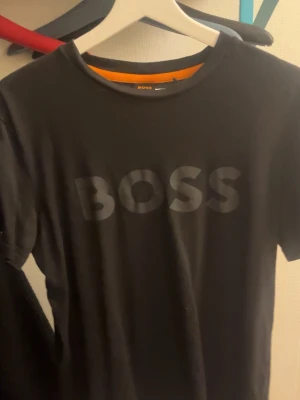 Svart BOSS t-shirt med tryck - Svart t-shirt från BOSS i storlek M med stort, ton-i-ton BOSS-tryck på bröstet. Klassisk rund hals och korta ärmar. Skön och mjuk känsla, perfekt för dig som gillar stilrena och enkla plagg med tydlig logga.
