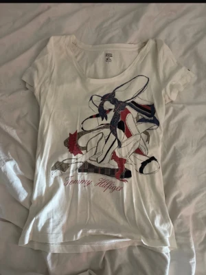 Vit t-shirt med tryck från Tommy Hilfiger - Snygg vit t-shirt från Tommy Hilfiger med ett stort grafiskt tryck i rött, blått och svart framtill. Klassisk rund halsringning och korta ärmar. Tillverkad i mjuk bomull som känns skön mot huden. 