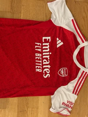 Arsenal matchtröja röd/vit - Säljer en Arsenal matchtröja från Adidas i rött och vitt med tryck på bröstet och ärmarna. Tröjan har vita detaljer, klubbmärke och sponsorlogga framtill samt namn och nummer 14 på ryggen. Tillverkad i lätt och ventilerande polyester, perfekt för fotboll. Aldrig använd
