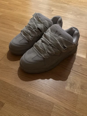 Gråa bershka skor - Säljer ett par grå chunky sneakers med breda, platta snören och rund tå. Skorna har en tjock sula och ovandel i syntetmaterial och mockaimitation. Perfekta för dig som gillar streetwear och vill sticka ut med detaljerade snörningar.
