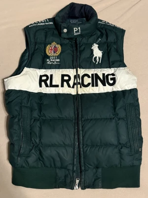 Grön dunväst från Ralph Lauren RL Racing - Säljer en grön dunväst från Ralph Lauren RL Racing med vit panel och stora broderade loggor fram och bak. Västen har dragkedja, ribbade muddar och coola racingdetaljer som broderad flagga och siffran 8 på ryggen. Perfekt för dig som gillar sportig stil.