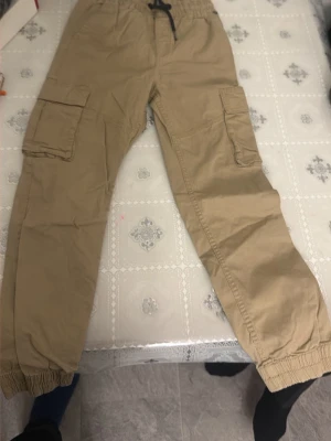 Beiga cargopants med resår - Säljer ett par beiga cargopants med resår i midjan och bensluten. Byxorna har stora sidofickor och snörning i midjan för extra komfort. Perfekta för en avslappnad och trendig look.