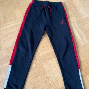 Jordan mjukisbyxor svart/röd/vit - Säljer ett par mjukisbyxor från Jordan i svart med röda detaljer och klassisk Air Jordan-logga på benet. Byxorna har dragsko i midjan och är tillverkade i mjukt bomullsmaterial, perfekta för chill eller sport. Passar dig som gillar streetwear och sportig stil. Stl 13-15 år, ca 160 cl