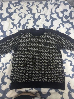 Svart EA7 Emporio Armani sweatshirt - Säljer en svart sweatshirt från EA7 Emporio Armani med all-over logomönster i vitt. Tröjan har rund hals, långa ärmar och ribbade muddar. Tillverkad i mjuk bomull för skön känsla och snygg streetstil.