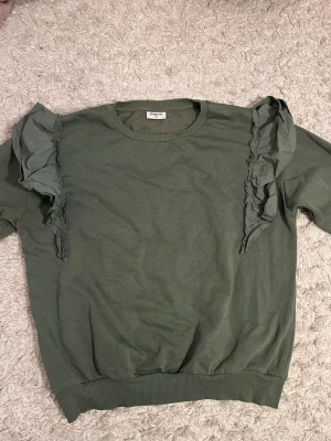 Grön sweatshirt med volang från Kappahl - Snygg grön sweatshirt från Kappahl i storlek 170. Tröjan har långa ärmar, ribbade muddar och coola volangdetaljer längs axlarna som ger en unik look. 💞