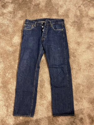 Levi's 501 mörkblå jeans W32 L30 - Klassiska Levi's 501 jeans i mörkblå tvätt med raka ben och knappgylf. Snygga kontrastsömmar och ikonisk läderpatch bak. Jeansen har fem fickor och är tillverkade i slitstarkt bomullsjeansmaterial. Perfekt för dig som gillar tidlös stil och vill ha ett par jeans som alltid funkar.