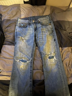 True Religion blå slitna jeans - Säljer ett par blå jeans från True Religion med slitningar och hål på knäna. Klassisk kontrastsöm och stora bakfickor med lock och tryckknapp. Jeansen har normal passform och är tillverkade i bomull med en snygg tvättad look.