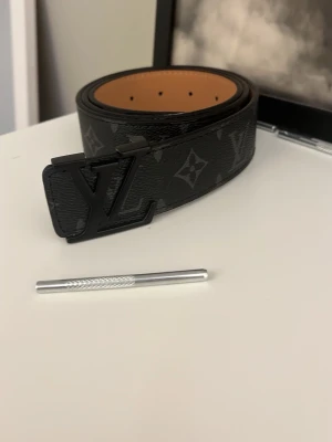 L&V  bälte svart - Säljer ett  bälte i svart med klassiskt monogrammönster och stor -logga i matt svart metall. Bältet är gjort i skinn med brun insida och har exklusiv känsla. Perfekt accessoar för att lyfta vilken outfit som helst.