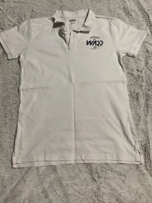 Vit polotopp från Warp med tryck - Säljer en vit polotopp från Warp med korta ärmar och klassisk krage. Toppen har två knappar vid halsen och ett snyggt mörkblått tryck på bröstet med texten 'Hardgoods EST 1996'. Materialet är mjuk bomull som känns skönt mot huden.