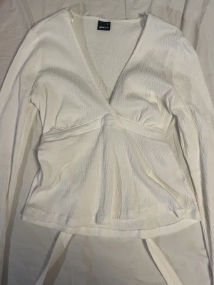 Vit omlottopp från Gina Tricot - Säljer en vit omlottopp från Gina Tricot med lång ärm och v-ringad hals. Toppen har en lätt struktur i tyget och knytband i midjan för en snygg passform. Perfekt att styla med jeans eller kjol för en fräsch look.