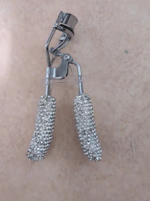 Ögonfransböjare - Säljer en silverfärgad ögonfransböjare med handtag täckta av glittrande strass. Metallkroppen ger stabilitet och handtagen är dekorerade med små, klara stenar för en lyxig känsla. Perfekt för att få snygg böj på fransarna och addera lite extra glam till sminkrutinen.