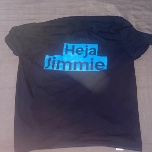 Svart t-shirt med blå text - Svart t-shirt med trycket 'Heja Jimmie' i stor, blå text på bröstet. Klassisk passform med korta ärmar och rund hals. Perfekt för dig som vill sticka ut med ett tydligt statement. Materialet känns mjukt och bekvämt mot huden.