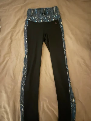 Svarta leggings från Drop of Mindfulness - Snygga svarta leggings från Drop of Mindfulness med bred, högmidjad linning och blått mönster längs sidorna och midjan. Tillverkade i stretchigt material med 86% polyester och 14% elastan, perfekta för träning eller chill. Mönstret har inslag av blått, vitt och grått.