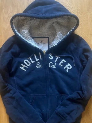 Hollister zip up med ull - Mjuk och varm Hollister zip-hoodie i mörkblå färg. Fodrad med teddyfleece på insidan – perfekt för kyliga dagar. Snygg broderad logga framtill.  Info: • Märke: Hollister • Storlek: S • Färg: Mörkblå • Modell: Zip-hoodie med huva • Skick: Bra begagnat skick