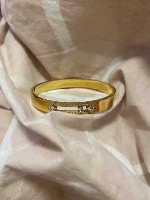 Guldfärgat armband med stenar - Snyggt guldfärgat armband med glittrande stenar framtill. Armbandet har en stilren och modern design med två större stenar i mitten och små stenar runtom. Perfekt accessoar för att lyfta din outfit.