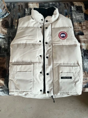 Beige dunväst från Canada Goose - Snygg beige dunväst från Canada Goose med hög krage, svarta knappar och dragkedja. Västen har två stora fickor framtill och klassisk logga på bröstet. Perfekt för lager-på-lager och håller dig varm under kyliga dagar.