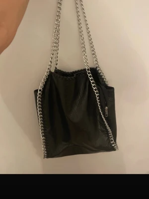 Svart axelväska med kedja från Tiamo - Snygg svart axelväska från Tiamo i fejkskinn med grova silverfärgade kedjor som axelband. Väskan har dragkedja på utsidan och detaljerad kant med flätad söm. Perfekt storlek för att få plats med allt du behöver och ger en cool vibe till din outfit.