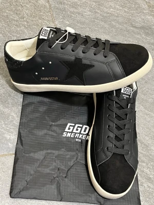 Svarta GGDB sneakers med stjärndetalj - Snygga svarta sneakers från GGDB med svart stjärna på sidan, vita detaljer och svart glittrig häl. Skorna har snörning, rund tå och vit platt sula. Ovandelen är i skinn och mocka, vilket ger en lyxig känsla. Perfekta för dig som vill ha en clean men ändå unik look.