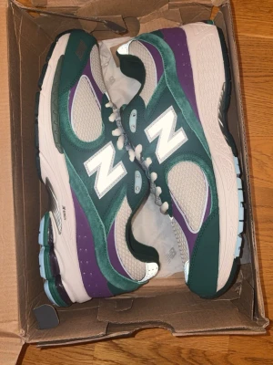 New Balance sneakers grön/lila mesh - Säljer ett par New Balance sneakers med grön och lila ovandel i mesh och mocka, vita detaljer och snörning. Sulan är vit med inslag av ljusblått och svart, och skorna har en stor vit N-logga på sidan. Perfekta för dig som gillar färg och sportig stil.