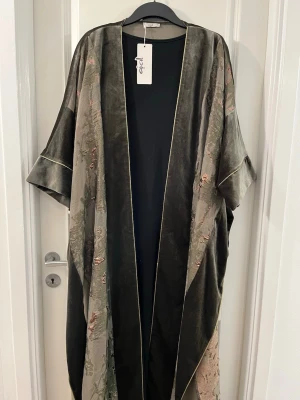 Grå sammet Kaftan/Abaya från Ajal - Säljer en unik grå Kaftan/Abaya från Ajal i mjuk sammet med breda, lösa ärmar och subtila broderade detaljer i rosa och grönt. Koftan har öppen front och tunna ljusa kantlinjer som ger en cool touch. Perfekt för dig som gillar statement-plagg.