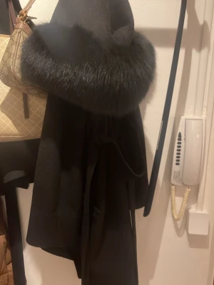 Svart kappa med päls från Hollies - Säljer en svart kappa från Hollies med stor fluffig päls runt kragen. Kappan har en elegant och klassisk form, med bälte i midjan för snygg passform. Materialet är mjukt och känns lyxigt, perfekt för kyliga dagar. Längst upp har ena knappen gått av men inget man märker av eller om man syr dit en egen. 