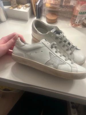 GGDB Superstar sneakers med glitterstjärna - Säljer ett par GGDB Superstar sneakers i vitt skinn med silverfärgade glitterdetaljer på hälen och en glittrig stjärna på sidan. Skorna har grå snörning och svart/vit etikett på plösen. Snygg platt sula i gummi och rund tå.