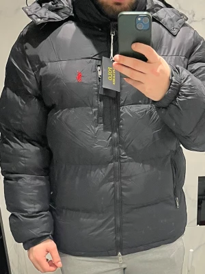 Svart pufferjacka från Polo Ralph Lauren - Svart pufferjacka från Polo Ralph Lauren med röd logga på bröstet. Jackan har huva, dragkedja framtill och två sidofickor med dragkedja. Materialet är polyester och jackan har en quiltad design som ger en schysst volym. Perfekt för kalla dagar.
