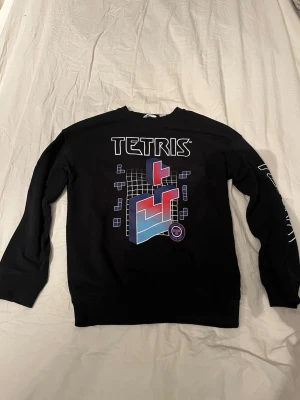 Svart Tetris sweatshirt med tryck från H&M  - Cool svart sweatshirt med stort Tetris-tryck framtill i blått, rött och vitt. Tetris-logga även på ena ärmen. Tröjan har rund hals och långa ärmar med muddar. Perfekt för dig som gillar retrospel och streetstyle.