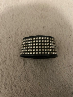 Svart skinnarmband med nitar - Coolt svart armband i skinn med flera rader silvriga nitar som ger en edgy vibe. Armbandet har tryckknappar för enkel stängning och passar perfekt till en rockig stil. Materialet är skinn och detaljerna är i metall. Snygg accessoar. Tyvärr lite skinn som gått up, men syns inte.