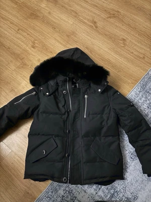 Svart Moose Knuckles dunjacka S - Svart dunjacka från Moose Knuckles med fet broderad hockeypatch på insidan. Jackan har pufferstil, dragkedja, knappstängning och fickor med lock. Huva med svart fuskpäls och coola detaljer som metallöglor och innerfickor. Perfekt för kalla dagar.