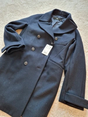 Mörkblå ullkappa från Uniqlo +J - Snygg mörkblå kappa från Uniqlo +J med dubbelknäppning och bred krage. Tillverkad i 100% ull med foder i polyamid och polyester. Kappan har långa ärmar, fickor och en clean, klassisk look som funkar till alla outfits.