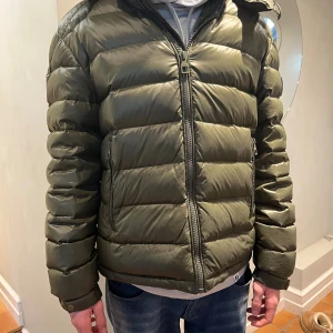 Olivgrön pufferjacka från Bvaldi - Olivgrön pufferjacka från Bvaldi med huva och  logomärke på ärmen. Fickor och en innerficka med dragkedja. Man kan reglera och dra åt linningen längst ner på i sidan.  Storlek M i Bvaldis storlek. Killen på bilden är 180cm lång och har stl s i andra klädesplagg. Mycket fint skick! Nypris: 3000kr