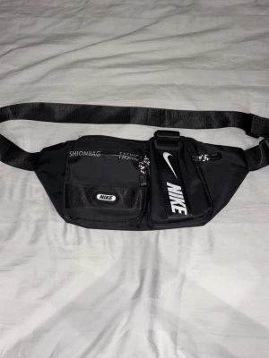 Svart midjeväska från Nike - Svart midjeväska från Nike med justerbart band och flera fack med dragkedja. Väskan har tydliga Nike-loggor i vitt på framsidan och är tillverkad i slitstark polyester. Perfekt för att bära med sig det viktigaste och hålla händerna fria.