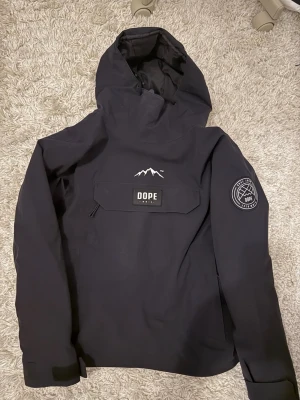 Svart skid/snowboardjacka Dope  - Säljer en svart snowboardjacka från Dope, modell Blizzard W. Jackan är riktigt praktisk då den har huva som man kan dra över hjälmen, och dragkedjor på sidorna som man kan öppna upp ifall man är varm! Den är även så gott så ny utan några tecken på användning. Normal pris är 2099kr