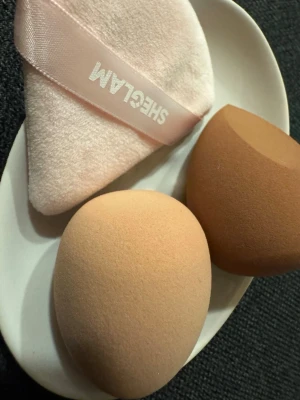 SHEGLAM  puff - Säljer två mjuka makeupsvampar i beige och brun samt en trekantig puderpuff i ljusrosa med SHEGLAM-logga. Perfekta för att applicera foundation, concealer eller puder. Puffen har ett praktiskt band för grepp och svamparna har rundad och snedskuren form för enkel applicering.