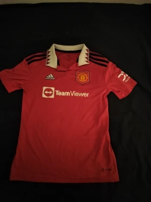Röd Manchester United Adidas tröja - Säljer en röd Manchester United tröja från Adidas med vit krage och svarta detaljer. Tröjan har klubbmärket broderat på bröstet och TeamViewer-logga framtill. Kragen har ett unikt mönster i rött och svart. Perfekt för fotbollsfans!