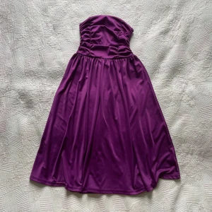 Lila axelbandslös långklänning H&M - Säljer en snygg lila axelbandslös långklänning från H&M. Klänningen har en glansig satinliknande yta, rynkad överdel och en vid, svepande kjol. Dragkedja i ryggen för enkel påklädning. Perfekt för dig som vill sticka ut med färg. Storlek 34 och använd ett fåtal gånger.