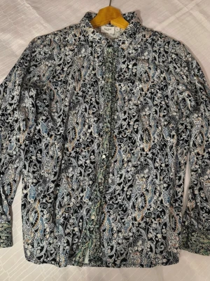 Mönstrad skjorta från Saint XS - Säljer en långärmad skjorta från Saint med detaljerat paisleymönster i svart, blått, grönt och vitt. Skjortan har knappar framtill och vid manschetterna, samt klassisk krage. Materialet känns som bomull och har en mjuk känsla. Perfekt för dig som gillar unika prints.