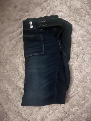 Mörkblå jeans från Gina Tricot - Snygga mörkblå jeans från Gina Tricot med två silverfärgade knappar i midjan och klassiska fickor fram. Jeansen har kontrastsömmar och är tillverkade i jeansmaterial. Perfekt för dig som gillar en enkel och stilren look.