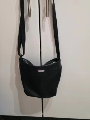 Svart axelväska från Don Donna - Säljer en stilren svart axelväska från Don Donna i skinnimitation. Väskan har en bred axelrem, silverfärgad logga framtill och praktisk dragkedja både upptill och på baksidan. Perfekt storlek för att få plats med allt nödvändigt och passar till många olika outfits.