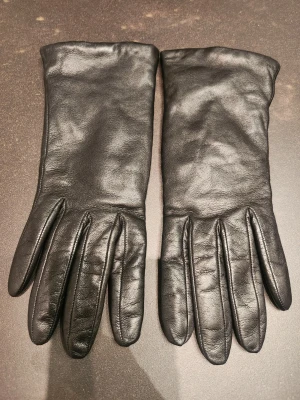 Svarta skinnhandskar storlek 7,5 - Snygga svarta handskar i mjukt skinn med klassisk design och diskreta sömmar. Insidan är fodrad med cashmere för extra värme och komfort. Eleganta och varma