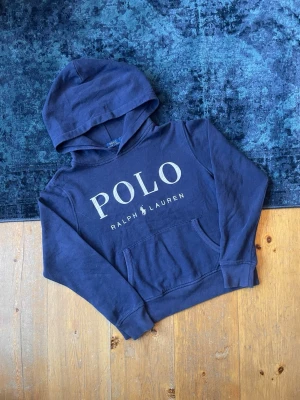 Mörkblå Ralph Lauren Hoodie  - Hej! Säljer en snygg, mörkblå Ralph Lauren hoodie med ”POLO” text på bröstet. Texten på framsidan är lite skadad (se bild), men det syns knappt vid användning. Storleken är M i barnstorlek, men skulle tro att den passar runt xxs. Hör av er vid frågor!